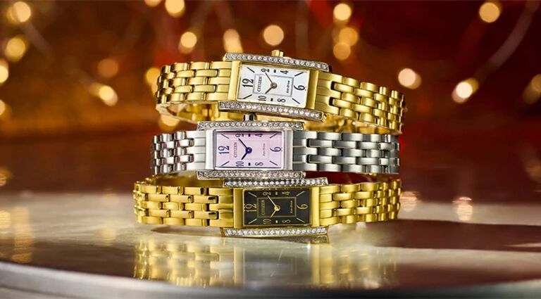 Women's Rectangular-Faced Watches; Image of three Petite Palidoro watch models EG7110-54Y, EG7112-59E and EG7112-59D on a table with holiday lights in the background