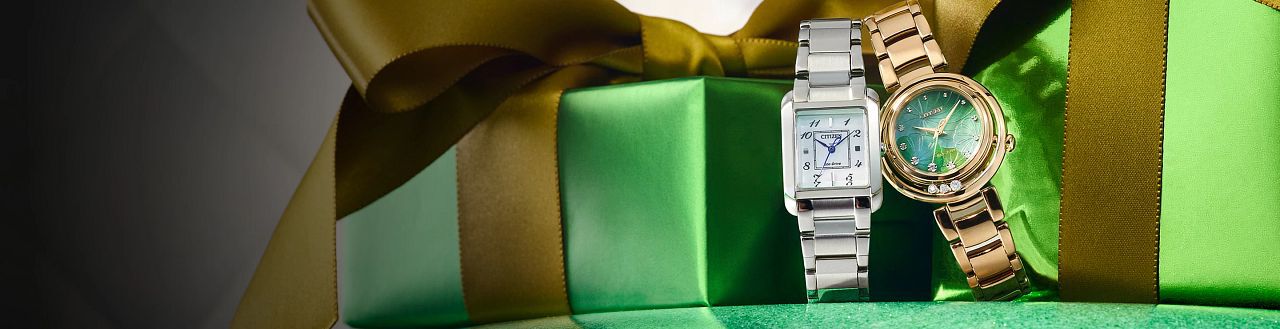 Image of two Citizen L watches sitting in front of green holiday gift boxes. Featuring Citizen L Bianca watch model EW5600-52D and Citizen L Arcly watch model EM1113-58Y