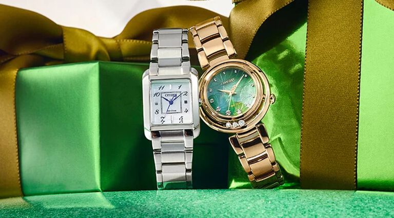 Image of two Citizen L watches sitting in front of green holiday gift boxes. Featuring Citizen L Bianca watch model EW5600-52D and Citizen L Arcly watch model EM1113-58Y